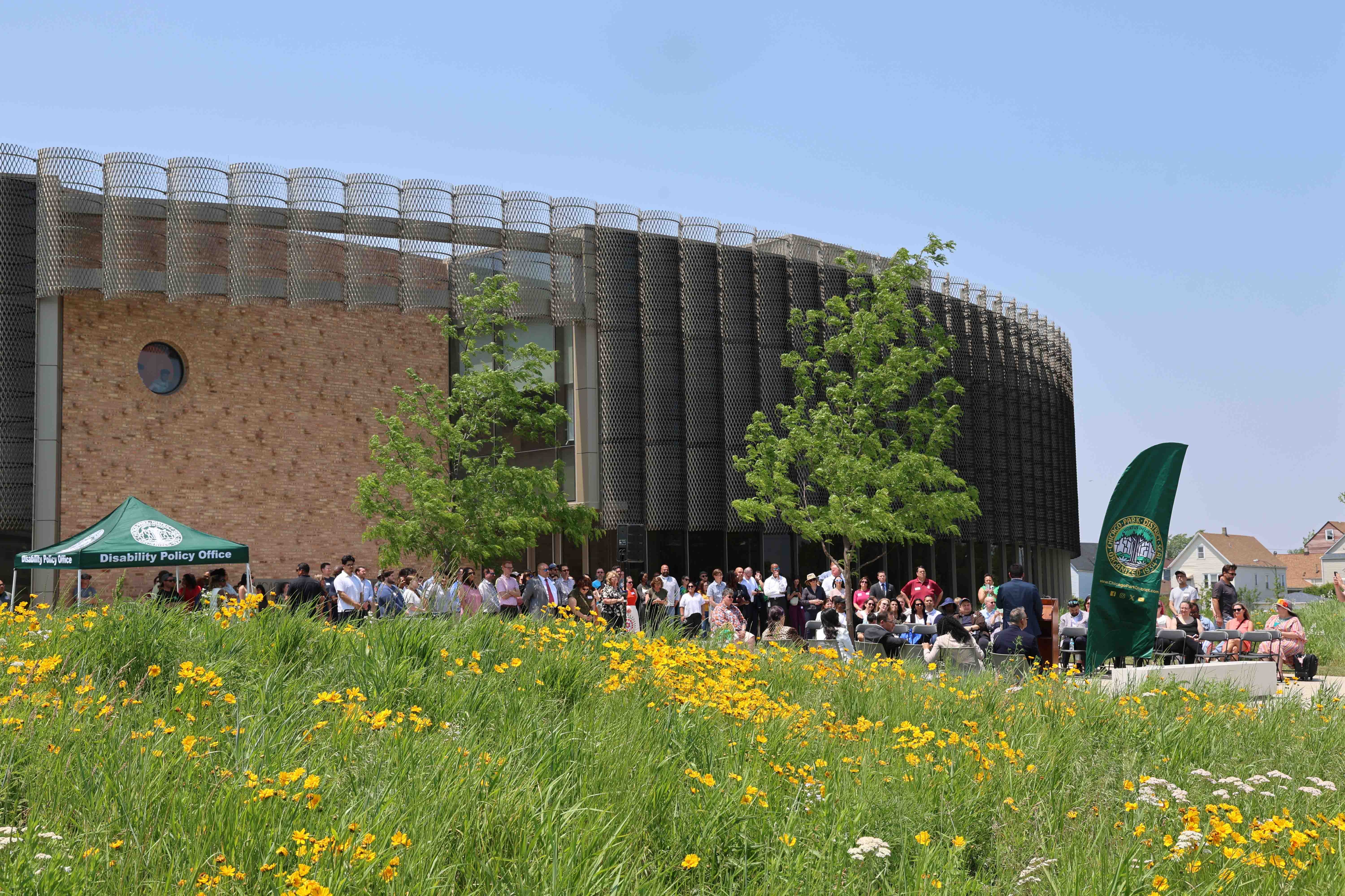 Crowd gathers outside a modern brick and metal building, with a field of yellow wildflowers in the foreground.